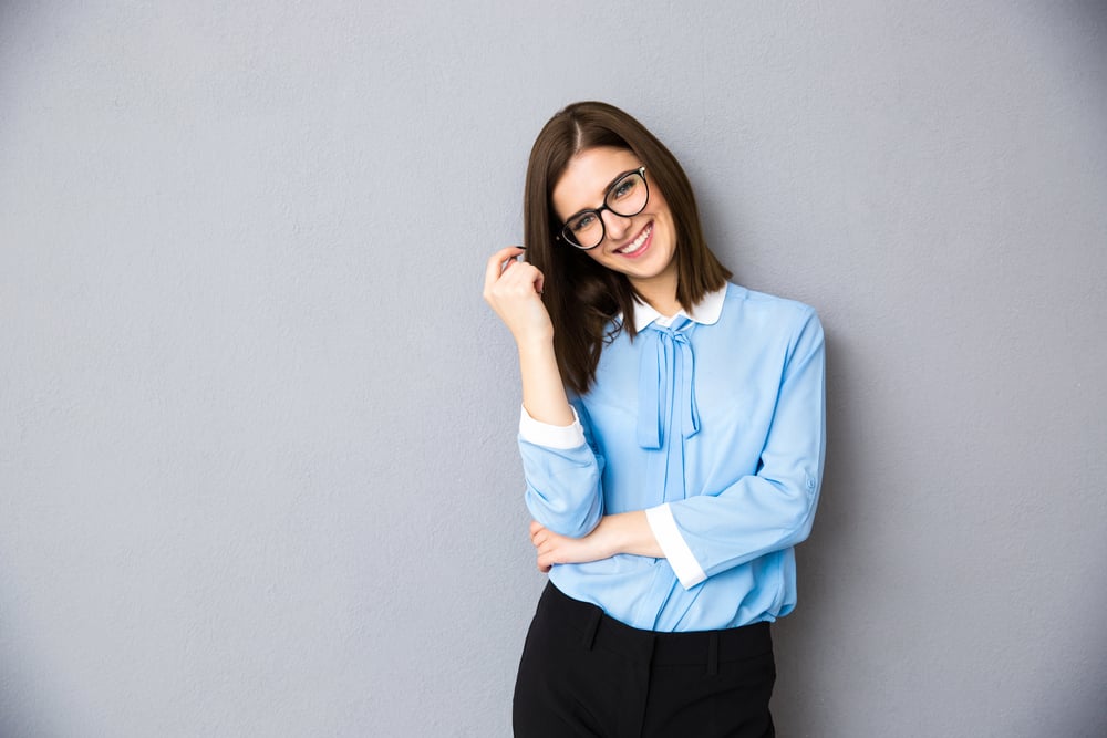 Smiling businesswoman in glasses standing over gray background. Wearing in blue shirt and glasses. Looking at camera-1