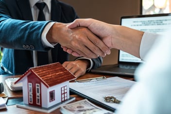 Two people shaking hands over a desk with a small house, contract, keys, and cash